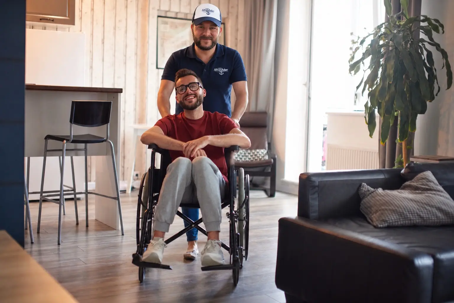Maximilian Schulze mit AlltagsAnker-Cap schiebt einen lächelnden jungen Mann im Rollstuhl durch dessen Wohnung